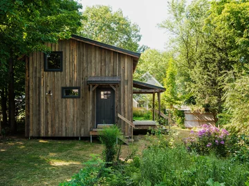 A finished New Frameworks Casita on its site, board-sided and weathered, with a covered porch and planting up to the steps
